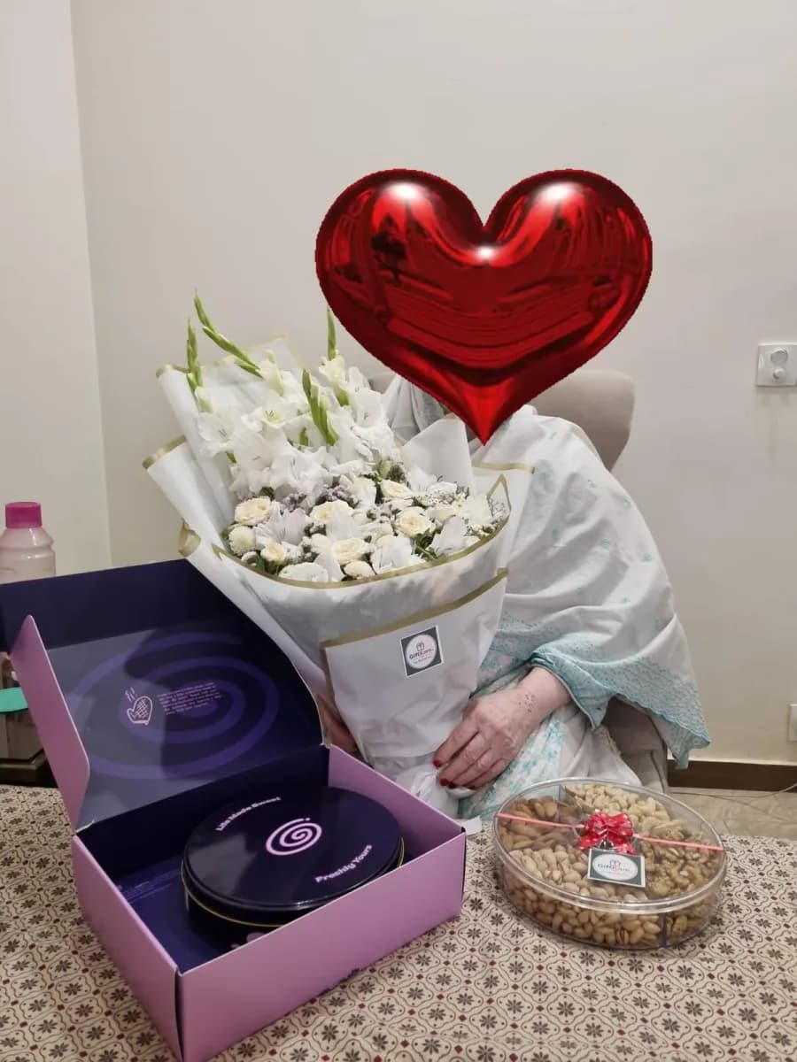 Customer holding a white rose and gladiolus bouquet beside a GiftKaro mithai box and a tray of dry fruit